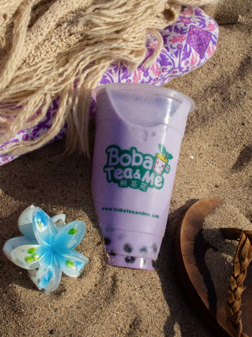 A purple Boba Tea & Me drink on beach sand beside sandals and a flower.